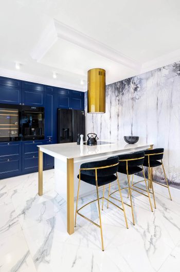 Blat kuchenny wykonany ze spieku kwarcowego Marazzi ''Statuario'' lux. Przy wyspie trzy czarno-złote krzesła.