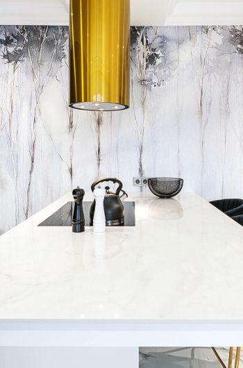 Wyspa kuchenna ze spieku kwarcowego Marazzi ''Statuario'' lux. Nad wyspą złoty, w kształcie tuby okap.