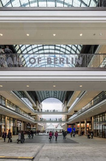 Na zdjęciu widzimy wnętrze centrum handlowego Mall of Berlin – nowoczesnej galerii zakupowej zlokalizowanej w samym sercu Berlina. Głównym elementem kompozycji jest szeroki, zadaszony pasaż, otoczony po bokach dwupoziomowymi galeriami handlowymi. Przez środek przebiega przeszklony dach w kształcie łuku, który doświetla przestrzeń naturalnym światłem, nadając całości przestronności i lekkości. Na balustradzie jednego z górnych poziomów umieszczony jest napis MALL OF BERLIN.
