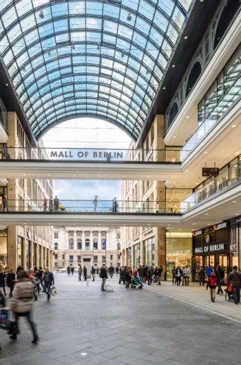 Zdjęcie przedstawia wnętrze centrum handlowego Mall of Berlin. Widać szeroką aleję z licznymi sklepami po obu stronach, takimi jak Zara, MAC, Saturn i inne. Na górze znajduje się przeszklony dach, który wpuszcza dużo naturalnego światła. Na pierwszym piętrze widać mostek łączący obie strony centrum handlowego, na którym znajduje się napis 
