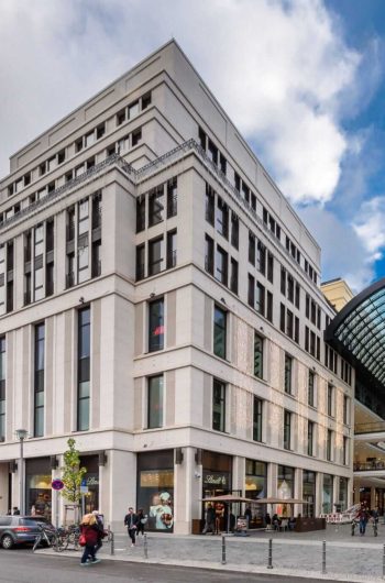 Zdjęcie przedstawia nowoczesną miejską scenerię z dużym kompleksem budynków, który posiada przeszklone atrium. Budynek ma kilka pięter i wyróżnia się współczesnym stylem architektonicznym z czystymi liniami i dużymi oknami. Atrium łączy dwie części budynku i jest pokryte zakrzywionym szklanym dachem, który wpuszcza naturalne światło, rozjaśniając przestrzeń poniżej.