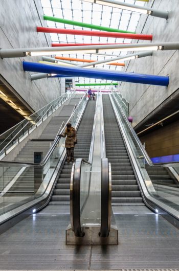 Na tym zdjęciu schody ruchome pokazane z dołu. Nad schodami kolorowe rury, w których jest oświetlenie. Schody i ściany na stacji metra w Lipsku zostały wykonane z kamienia naturalnego.
