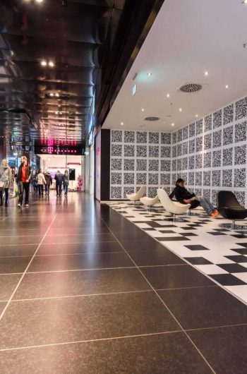 Zdjęcie przedstawia wnętrze nowoczesnej przestrzeni handlowej „Höfe am Brühl” w Lipsku. Podłoga wyłożona jest dużymi, ciemnymi płytkami, a sufit ma połyskujące, metaliczne wykończenie z wbudowanym oświetleniem. Po lewej stronie widać kilka osób spacerujących – jedna z nich niesie torby z zakupami. Po prawej znajduje się strefa wypoczynkowa z designerskimi krzesłami i ciekawą ścianą pokrytą kodami QR. Nad nimi zamontowano ekrany, na których wyświetlany jest czerwony obiekt na tle błękitnego nieba.