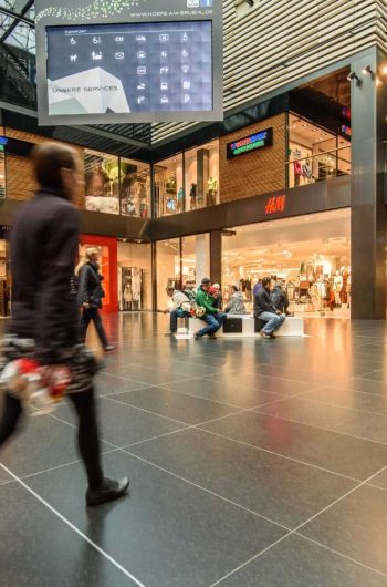 Na zdjęciu widzimy wnętrze nowoczesnej galerii handlowej „Höfe am Brühl” w Lipsku. Przestrzeń jest jasna i przestronna, z elegancką posadzką i elementami ze szkła. Po lewej stronie zauważalne są sklepy takie jak New Yorker i H&M, a w centrum znajduje się duży cyfrowy ekran z napisem „UNSERE SERVICES” oraz ikonami przedstawiającymi dostępne udogodnienia. Widać ludzi spacerujących i odpoczywających na ławkach.