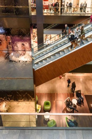Na zdjęciu widzimy wnętrze wielopoziomowego centrum handlowego „Höfe am Brühl” w Lipsku o nowoczesnej architekturze. Centralnym punktem kompozycji jest ruchoma schodnia, po której przemieszczają się ludzie między piętrami. Po jej lewej stronie znajduje się efektowny, błyszczący element – prawdopodobnie ściana wodna lub dekoracyjna instalacja z refleksującym światłem materiałem. Na niższym poziomie mieści się strefa wypoczynkowa z zielonymi fotelami i niewielkimi stolikami. W lewym dolnym rogu można dostrzec infopunkt z ekranem cyfrowym, a wokół widać przechodniów oraz klientów sklepu, którzy nadają przestrzeni pulsujące życie.