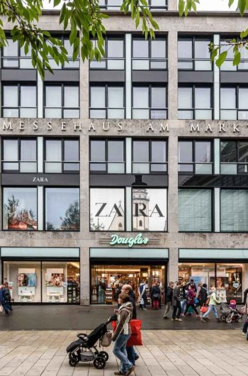 Na zdjęciu widzimy fasadę budynku Messehaus am Markt w Lipsku, sfotografowaną na wprost, dzięki czemu nazwa obiektu jest doskonale widoczna na górnej części elewacji. Charakterystyczna, nowoczesna architektura budynku opiera się na rytmicznym układzie dużych okien oraz jasnych, kamiennych ram konstrukcyjnych. Na parterze znajdują się liczne sklepy popularnych marek, takich jak Zara, Douglas, Vero Moda czy Jack&Jones. Witryny są jasno oświetlone, co przyciąga uwagę przechodniów. Przed Messehaus am Markt panuje ożywiony ruch – ludzie spacerują, robią zakupy, prowadzą wózki dziecięce lub mijają się w grupkach.