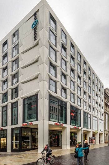 Na zdjęciu znajduje się nowoczesny, wielopiętrowy budynek o unikalnej geometrycznej fasadzie. Posiada duże okna, co nadaje mu przestronny wygląd. Budynek jest częścią przestrzeni miejskiej i ma na parterze sklepy oraz miejsca handlowe. Widać ludzi spacerujących i jeżdżących na rowerach na pierwszym planie. Pogoda wydaje się pochmurna, co nadaje zdjęciu stonowany klimat. Na rogu budynku widnieje szyld „Motel One”, wskazujący, że znajduje się tam hotel.