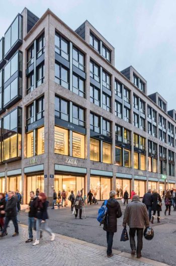 Na zdjęciu widać ruchliwe centrum Lipska, a głównym punktem kadru jest nowoczesny budynek Messehaus am Markt. Jego charakterystyczna fasada z dużymi przeszkleniami i regularnym układem okien tworzy efektowny, współczesny akcent w otoczeniu historycznych kamienic. Parter budynku wypełniają jasno oświetlone witryny znanych sklepów, co podkreśla handlowy charakter tego miejsca.