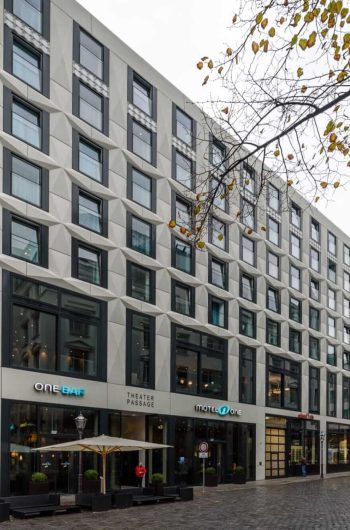 Na zdjęciu widzimy nowoczesną miejską ulicę, na której znajduje się kilka budynków o różnym stylu architektonicznym. Główny budynek w centrum ma współczesny design z geometryczną fasadą i dużymi oknami. W jego wnętrzu mieszczą się różne lokale, w tym „ONE BAR”, „THEATER PASSAGE” i „MOTEL ONE”. Po lewej stronie zdjęcia znajduje się starszy budynek z szyldem „ANTIQUARIAT”, sugerującym, że mieści się tam antykwariat. Ulica jest wyłożona brukiem, a przy niej rosną drzewa z jesiennymi liśćmi.