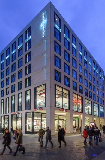 Na zdjęciu widać budynek, w którym znajduje się Motel One w Lipsku. Budynek z wieloma piętrami, który ma elegancki, współczesny design. Jego fasada jest podświetlona na niebiesko, co nadaje mu charakterystyczny wygląd. Parter budynku mieści kilka sklepów, których duże przeszklone witryny prezentują różne produkty. Przed budynkiem na ulicy poruszają się ludzie. Niebo na zdjęciu jest ciemnoniebieskie, co świadczy o tym, że zdjęcie wykonano wieczorem lub nocą.