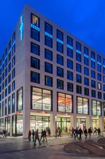 Na zdjęciu widzimy ulicę miejską w Lipsku w godzinach wieczornych. Głównym punktem obrazu jest nowoczesny, wielopiętrowy budynek z hotelem o nazwie „Motel One”, który znajduje się na rogu. Budynek ma duże okna i kilka sklepów na parterze, w tym „Shoe City” oraz „Nanu Nana”. Ulica jest pełna pieszych spacerujących. Niebo ma głęboki, ciemnoniebieski odcień, co wskazuje na późniejszą porę dnia.