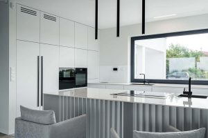 Na zdjęciu widoczna jest nowoczesna kuchnia w&nbsp;minimalistycznym stylu. Dominującym kolorem jest biel, która nadaje przestrzeni jasności i&nbsp;elegancji. Fronty szafek kuchennych są gładkie, bez widocznych uchwytów. Centralnym punktem jest wyspa kuchenna z&nbsp;blatem wykonanym ze spieku kwarcowego Dekton o&nbsp;jasnym wykończeniu, idealnie odbijającym światło. Z&nbsp;przodu wyspy widać pionowe dekoracyjne panele, które dodają subtelnej struktury.