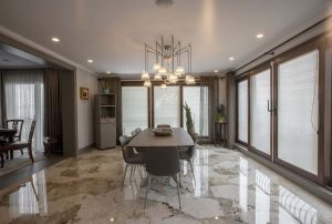 Zdjęcie przedstawia elegancką jadalnię urządzoną w nowoczesnym stylu. Centralnym punktem pomieszczenia jest prostokątny stół z ciemnym blatem, otoczony sześcioma minimalistycznymi krzesłami. Podłoga wyłożona jest luksusowymi płytkami w wysokim połysku, wykonana ze spieku kwarcowego Dekton, z wyraźnym użyłowaniem w odcieniach bieli i beżu. Pomieszczenie jest jasne dzięki dużym przeszklonym drzwiom balkonowym, które wpuszczają dużo naturalnego światła.