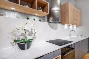 Zdjęcie przedstawia nowoczesny blat kuchenny o eleganckim i minimalistycznym designie. Blat jest biały wykonany z konglomeratu kwarcowego, a na nim znajduje się elektryczna płyta grzewcza z czterema palnikami. Nad płytą grzewczą wisi stalowy okap. Po prawej stronie płyty grzewczej znajduje się zlew z nowoczesnym, elastycznym kranem. Ściana za blatem, podobnie jak sam blat, wykonana z konglomeratu kwarcowego. Po lewej stronie blatu stoi czarna waza z białymi orchideami i zielonymi liśćmi. Nad blatem wiszą drewniane szafki z otwartymi półkami, na których znajdują się małe doniczki i szklane słoiki. Szafki posiadają oświetlenie podszafkowe, które oświetla blat. Całość projektu jest czysta, nowoczesna i estetycznie przyjemna.