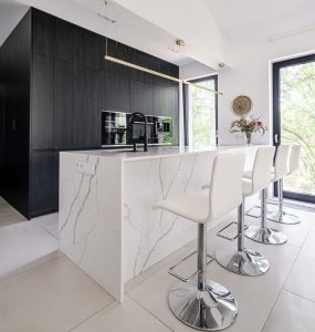 Na zdjęciu znajduje się nowoczesna kuchnia. Kuchnia ma wyspę kuchenną wykonaną z konglomeratu kwarcowego, przy której stoją cztery białe krzesła barowe z chromowanymi podstawami. Na wyspie znajduje się czarny kran i płyta grzewcza. W tle widać dużą szafę kuchenną w kolorze czarnym, w której zamontowane są dwa piekarniki. Kuchnia jest dobrze oświetlona dzięki dużym oknom, przez które widać zieleń na zewnątrz. Na ścianie obok okna wisi zegar, a na blacie stoi wazon z kwiatami.