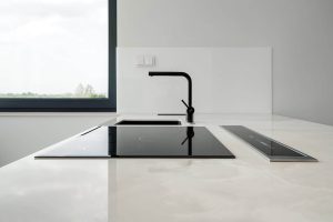 Zdjęcie przedstawia nowoczesny, minimalistyczny blat kuchenny o eleganckim designie. Blat wykonany jest z jasnego materiału, spieku kwarcowego Dekton. W blacie wbudowana jest czarna płyta indukcyjna z czterema palnikami. Po prawej stronie płyty grzewczej znajduje się długi, wąski, czarny panel, który wygląda jak system wentylacji wyciąganej w dół. W tle widać czarny kran z pojedynczym uchwytem, zainstalowany nad prostokątnym zlewem. Kuchnia ma duże okno z czarną ramą, które dostarcza naturalne światło i widok na zewnątrz.