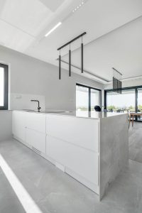 Zdjęcie przedstawia nowoczesne, minimalistyczne wnętrze kuchni. Kuchnia posiada dużą białą wyspę z blatem, wykonanym ze spieku kwarcowego Dekton. Nad wyspą wiszą trzy eleganckie, czarne lampy wiszące, które dodają całości stylowego wyglądu. Kuchnia ma duże okna, które wpuszczają naturalne światło, tworząc jasną i przestronną atmosferę. Podłoga jest w jasnoszarym kolorze, co doskonale komponuje się z ogólną kolorystyką kuchni. W tle znajduje się jadalnia z stołem i krzesłami oraz dużymi przesuwnymi drzwiami, które prowadzą na zewnątrz.