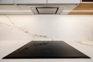 Obraz przedstawia nowoczesną kuchnię z elegancką płytą indukcyjną firmy Bosch, zamontowaną na białym blacie wykonanym ze spieku kwarcowego Neolith. . Nad płytą znajduje się stalowy okap wbudowany w zabudowę. Ściana za płytą jest również wyłożona ze spieku kwarcowego Neolith.