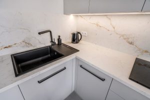 Zdjęcie przedstawia nowoczesną kuchnię z białym blatem i wykonanym ze spieku kwarcowego Neolith, który przechodzi również na ścianę między szafkami górnymi i blatem. Na pierwszym planie znajduje się czarny zlew z czarną baterią oraz dozownikiem na mydło. Obok zlewu stoi czajnik elektryczny. Na blacie po prawej stronie widoczna jest płyta indukcyjna