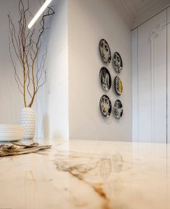 Obraz przedstawia nowoczesne wnętrze z blatem wykonanym ze spieku kwarcowego Neolith. Po lewej stronie blatu znajduje się biała waza o teksturowanym wzorze, w której są wysokie, cienkie gałązki. Obok wazy znajdują się białe miski oraz złożona serwetka. W tle widoczna jest ściana ozdobiona sześcioma dekoracyjnymi talerzami ułożonymi w dwóch pionowych kolumnach. Każdy talerz ma czarno-biały artystyczny wzór twarzy. Ściana po prawej stronie ma dekoracyjne zdobienia w formie sztukaterii.