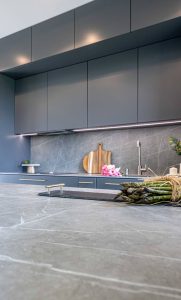 Na obrazku przedstawiona jest nowoczesna kuchnia. Widać w niej ciemnoszare szafki z dopasowanym blatem wykonanym ze spieku kwarcowego Neolith w tym samym odcieniu. Ściana między blatem a górnymi szafkami również została wykończona tym samym materiałem, tworząc spójną i elegancką całość.