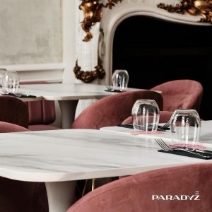 Obraz przedstawia stylowe wnętrze restauracji lub kawiarni, gdzie elegancja spotyka się z nowoczesnością. W centralnej części kompozycji znajdują się stoliki o białych, subtelnie połyskujących blatach wykonanych ze spieku kwarcowego Paradyż. Otoczone są przez aksamitne, różowe krzesła, które dodają wnętrzu przytulnego, ale jednocześnie luksusowego charakteru. Na stolikach ustawiono szklanki, starannie złożone serwetki oraz sztućce, umieszczone na czarnych serwetkach, co tworzy elegancki kontrast. W tle widać dekoracyjny kominek z białymi i złotymi akcentami, który podkreśla wyrafinowany styl pomieszczenia.