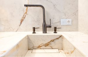 Na zdjęciu widoczny jest elegancki zlew, blat oraz okładzina ścienna wykonane ze spieku kwarcowego Neolith. Nad zlewem zamontowana została srebrna bateria o minimalistycznym designie, z dwoma pokrętłami po bokach. W tle znajduje się gniazdko elektryczne.