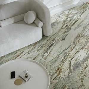 Obraz przedstawia nowoczesne wnętrze z jasną sofą o zaokrąglonych poduszkach oraz okrągłym, białym stolikiem kawowym. Podłoga wykonana jest ze spieku kwarcowego Paradyż, który zachwyca niepowtarzalnym wzorem. Kolory zieleni, bieli i brązu nadają przestrzeni elegancki, luksusowy wygląd. Na stoliku znajdują się czarny smartfon, mała kwadratowa taca z okrągłym złotym elementem oraz dyfuzor zapachowy z patyczkami.