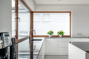 Na zdjęciu widać nowoczesną kuchnię o minimalistycznym wystroju. Dominuje w niej czystość i prostota — białe szafki, gładkie powierzchnie i dużo naturalnego światła wpadającego przez duże okna. Na blacie, tuż przy oknie, stoją dwie zielone rośliny doniczkowe, które dodają przytulnego akcentu tej eleganckiej przestrzeni.