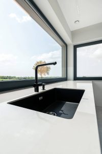 Na zdjęciu widzimy nowoczesny zlew kuchenny z czarnym, stylowym kranem i głęboką czarną komorą osadzoną w białym blacie. Minimalistyczny design podkreśla elegancję przestrzeni, a duże okno za zlewem wpuszcza sporo naturalnego światła, odsłaniając widok na zieleń i błękitne niebo.