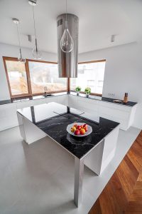Na zdjęciu znajduje się przestronna, nowoczesna kuchnia, która emanuje elegancją i funkcjonalnością. Centralnym punktem jest duża wyspa z czarnym blatem, wsparta na stalowych nogach, co dodaje industrialnego charakteru. Na blacie widoczna jest misa pełna owoców, wprowadzająca odrobinę koloru i świeżości do monochromatycznego wnętrza. Nad wyspą zawieszony jest okrągły stalowy okap z wbudowanym oświetleniem, który stanowi ciekawy, designerski element. Szafki kuchenne są białe i minimalistyczne, doskonale komponując się z jasnym światłem wpadającym przez duże okna. Na blacie obok okien ustawiono dwie zielone rośliny doniczkowe, które ożywiają przestrzeń. Podłoga jest połączeniem jasnych płytek i drewnianych paneli, co subtelnie dzieli strefy i dodaje wizualnej głębi.