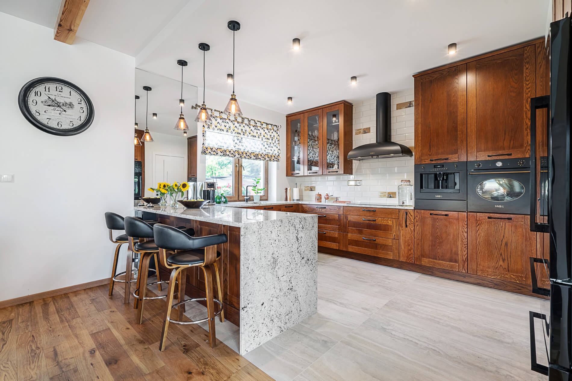 Na zdjęciu widoczna jest przestronna, klasyczno-nowoczesna kuchnia z&nbsp;wyraźnym akcentem naturalnego granitu. Jasny, nakrapiany granit został wykorzystany jako masywny blat wyspy kuchennej, nadając wnętrzu elegancji i&nbsp;solidności. Kamień kontrastuje z&nbsp;ciepłymi, drewnianymi frontami szafek oraz&nbsp;czarnymi detalami sprzętów AGD i&nbsp;oświetlenia.Wyspa pełni zarówno funkcję roboczą, jak i&nbsp;jadalnianą — ustawiono przy&nbsp;niej tapicerowane hokery. Granitowa powierzchnia prezentuje się szlachetnie i&nbsp;ponadczasowo, a&nbsp;jednocześnie podkreśla trwały, praktyczny charakter aranżacji.