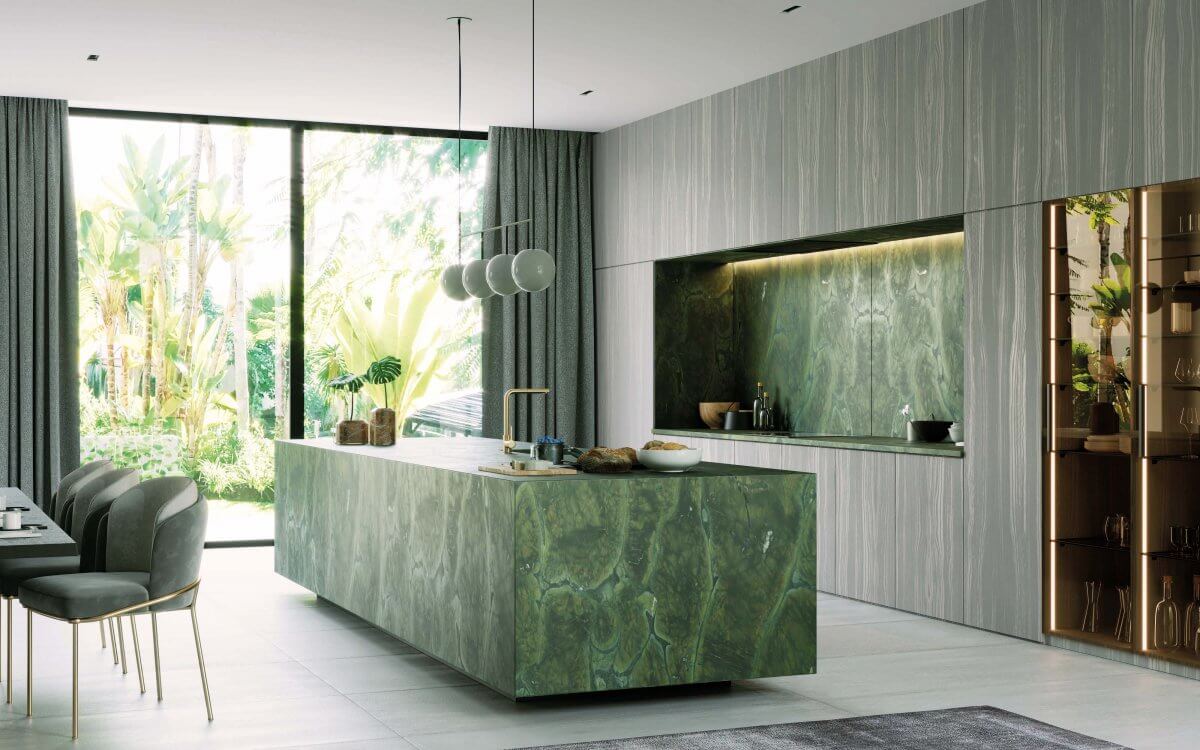 Na obrazie widoczna jest nowoczesna, minimalistyczna kuchnia, w której centralnym punktem aranżacji jest wyspa wykonana z zielonego kwarcytu. Kamień zachwyca głęboką, nasyconą barwą z wyraźnym, organicznym użyleniem, które przypomina naturalne struktury roślinne. Powierzchnia kwarcytu odbija światło wpadające przez duże przeszklenia, podkreślając jego wielowymiarową strukturę i subtelne przejścia tonalne.Ten sam materiał został zastosowany jako okładzina ścienna w strefie roboczej, tworząc spójną i elegancką kompozycję. Zielony kwarcyt kontrastuje z jasnymi, pionowo ryflowanymi frontami zabudowy oraz ciepłymi akcentami drewna i złotymi detalami armatury.