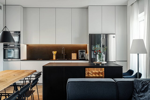 Na zdjęciu znajduje się nowoczesna kuchnia z&nbsp;otwartą strefą dzienną. Centralnym elementem jest wyspa kuchenna z&nbsp;ciemnym, granitowym blatem, który kontrastuje z&nbsp;jasną, minimalistyczną zabudową meblową. W&nbsp;tle widoczna jest pełna zabudowa ze sprzętem AGD oraz&nbsp;podświetlana przestrzeń robocza. Aranżację uzupełnia drewniany stół z&nbsp;czarnymi krzesłami oraz&nbsp;strefa wypoczynkowa z&nbsp;sofą, co tworzy spójną i&nbsp;elegancką przestrzeń.